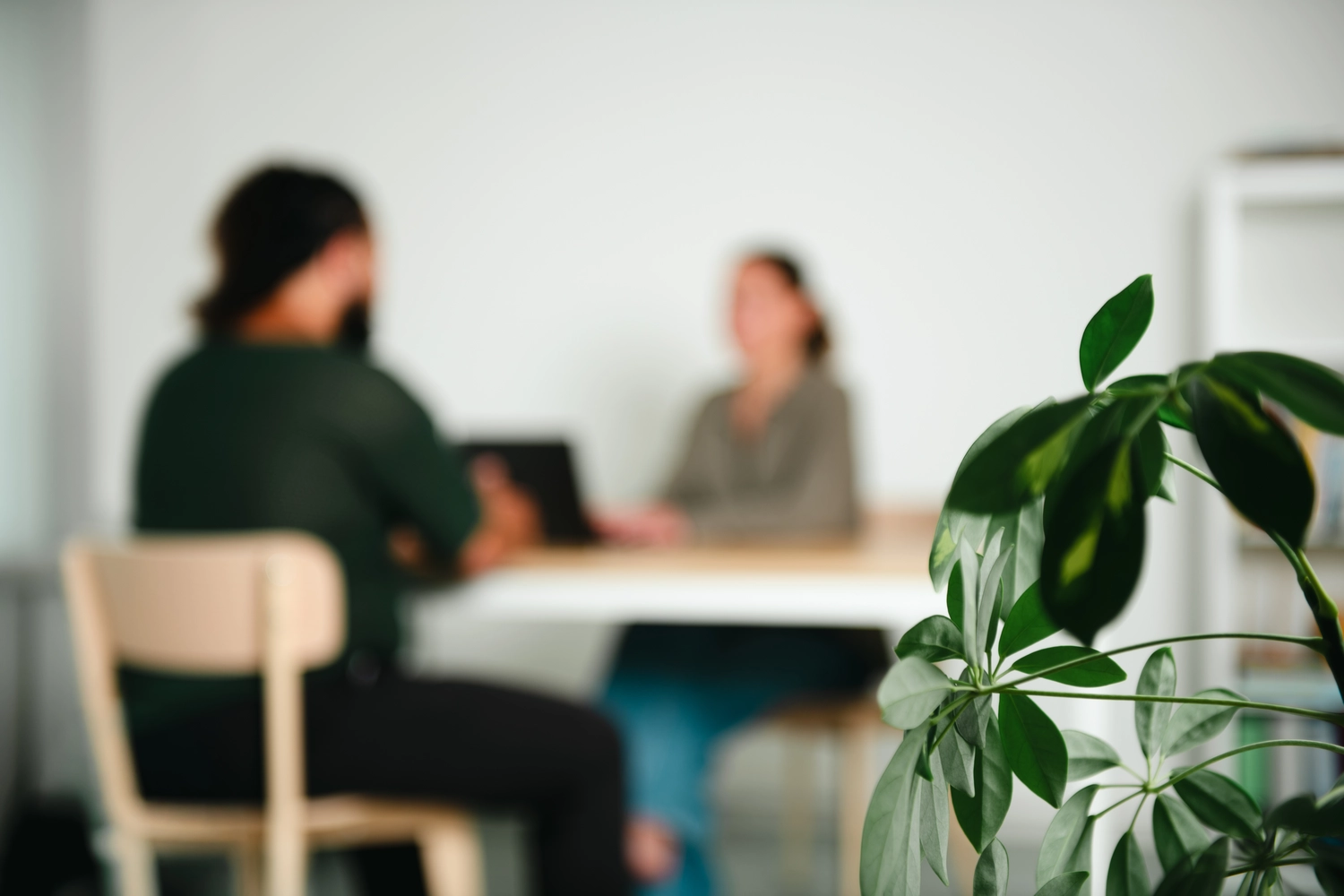 KONTEXT JUGEND – Rechtspsychologin – Im Vordergrund steht eine Pflanze im scharfen Fokus, während im Hintergrund zwei unscharfe Personen an einem Tisch mit einem Laptop sitzen.